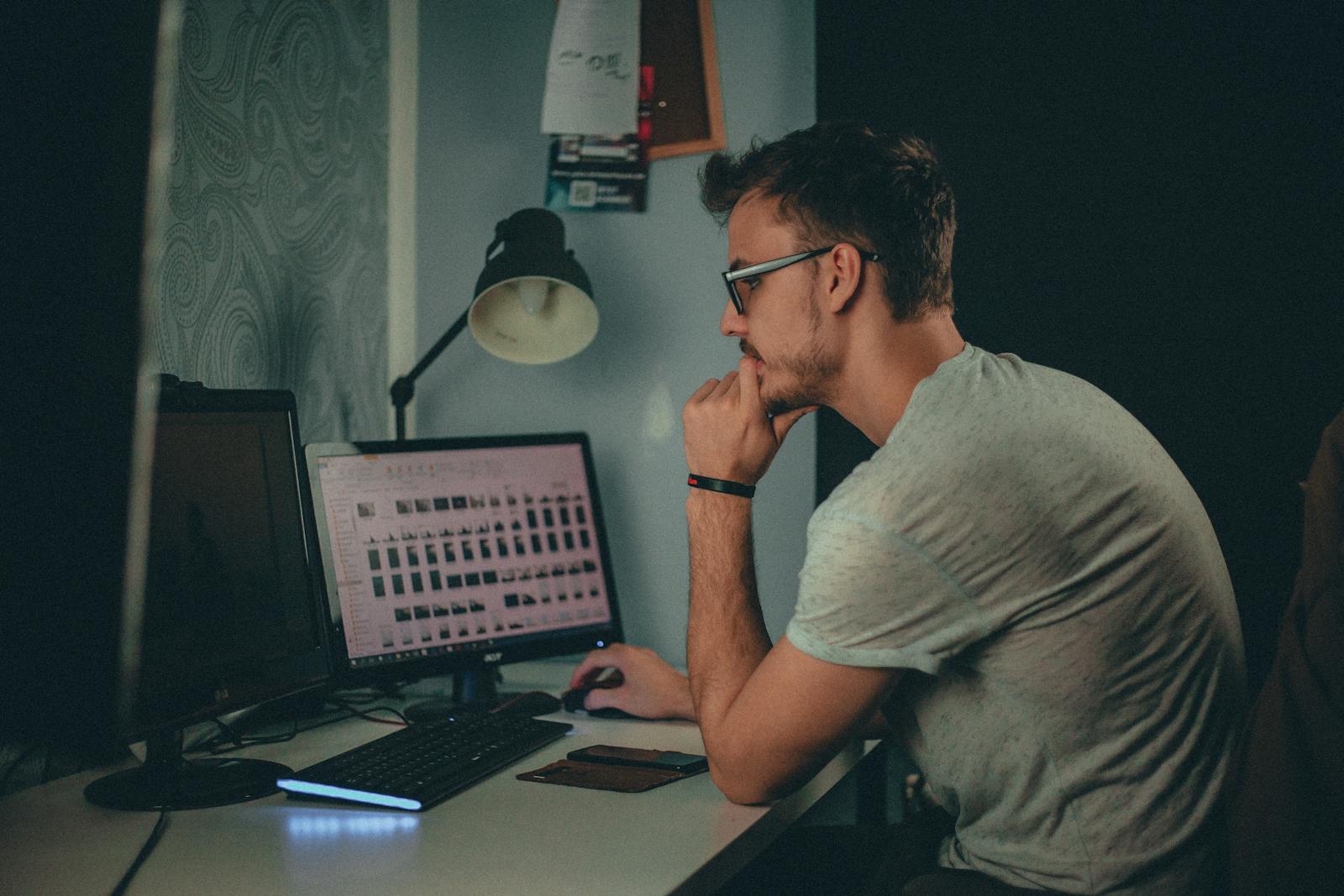 Focused man using a desktop computer in a dimly lit workspace with modern desk setup.